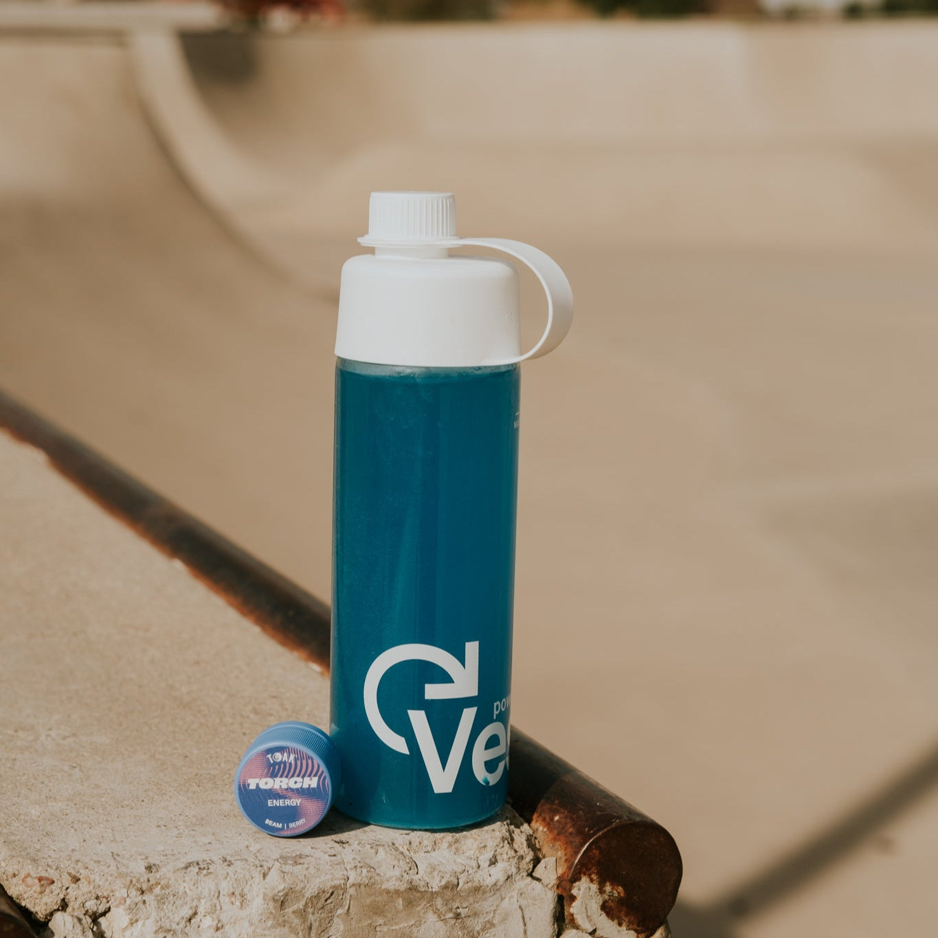 Blue water bottle with white cap and logo on a concrete surface at a skate park.